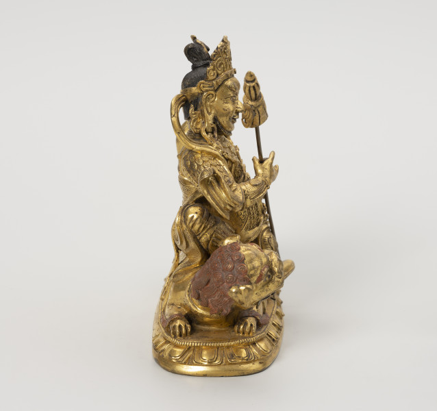 Wajśrawana (Dzambala) - ujęcie z lewej; Złota, męska postać siedząca na złotym, ryczącym lwie. W prawej ręce trzyma chorągiew zwycięstwa (osobny, wyciągany element). Postać w gniewnej formie ma upięte w kok czarne włosy, szlachetnie ubrana: nosi diadem, liczne ozdoby z klejnotów, a jej strój przypomina zbroję wysokiej rangi dostojnika/króla. Lwa o bordowych brwiach, sierści na głowie przedstawiono w pozycji wpółleżącej - waruje na ugiętych łapach, jest osiodłany. Obie postacie ustawiono na lotosowej podstawie. Posążek najprawdopodobniej przedstawia bóstwo bogactwa Wajśrawanę wraz z jego atrybutami.