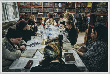 scena rodzajowa - Poziomy, barwny kadr fotograficzny przedstawiający grupę kobiet i dziewczynek siedzących przy długim stole. U szczytu stołu siedzi także dwóch mężczyzn z brodami, długimi pasmami włosów zakrywających uszy (pejsami) oraz w dużych futrzanych czapach w kształcie walca. Obok mężczyzn stoi dwoje małych dzieci. Zebrani pogrążeni są w czytaniu tekstów leżących przed każdą z osób, ich pozy wskazują na skupienie. Jedna z kobiet czyta i karmi butelką niemowlę leżące na jej kolanach. Stół stoi po środku pomieszczenia, którego ściany są zastawione w całości regałami, na których stoją książki w dekoracyjnych obwolutach (biblioteka?). Ścianę po lewej tronie kadru w całości wypełnia zasłonięte okno. Na stole nakrytym obrusem i przezroczystą ceratą stoją w części opróżnione butelki po napojach oraz rozpakowane folie i puste talerze. w pomieszczeniu pełnym regałów z książkami.