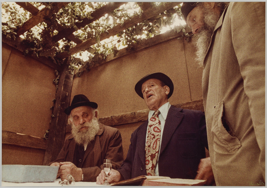 fotografia - Zdjęcie przedstawia trzech mężczyzn. Dwóch z nich siedzi przy stole (po lewej stronie kadru), trzeci stoi obok stołu (po prawej stronie kadru). Pierwszy mężczyzna od lewej ma na sobie brązowy płaszcz i czarny kapelusz. Ma siwą brodę. Zgięte w łokciach ręce opiera na stole. Drugi od lewej mężczyzna ma na sobie ciemny garnitur, jasną koszulę, wzorzysty krawat oraz ciemny kapelusz. Jest gładko ogolony. Prawą rękę opiera na stole. Trzyma w niej kieliszek wódki. Mężczyzna stojący przy stole widoczny jest z profilu. Ma na sobie jasną (beżową?) kurtkę/płaszcz oraz ciemne nakrycie głowy (czapkę?). Również ma siwą brodę. Oczy mężczyzn po prawej i lewej stronie są skierowane na kieliszek wódki. Mężczyźni znajdują się w prowizorycznym pomieszczeniu (szałasie). Za nimi widać ściany z płyty. Nad ich głowami widoczne ażurowe zadaszenie wykonane z listew ozdobionych liśćmi. 