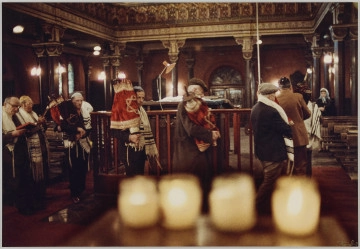 MPOLIN-M1598 fotografia - Zdjęcie przedstawia grupę mężczyzn we wnętrzu Synagogi. Część z nich obchodzi bimę trzymając w rękach zwoje tory w ozdobnych sukienkach. Po lewej stronie kadru widać dwóch mężczyzn trzymających w rękach otwarte księgi. Modlą się. Wszyscy mężczyźni mają nakrycia głowy (jarmułki lub kaszkiety). Ramiona większości z nich okrywają tałesy. Po prawej stronie kadru, w głębi synagogi widać modlącą się kobietę w błękitnej chuście na głowie. U dołu kadru widoczne cztery palące się świece. Świece są rozmazane, ostrość jest skupiona na mężczyznach.