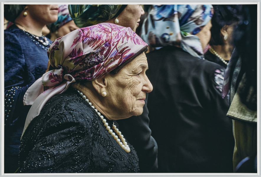 scena rodzajowa - Poziomy, barwny kadr fotograficzny przedstawiający na pierwszym planie postać starszej kobiety w czarnej sukience, białych perłach na szyi oraz wzorzystej chustce w odcieniach fioletu na głowie. Kobieta patrzy przed siebie, na drugim planie widać grupę podobnie ubranych kobiet, których postaci zostały ucięte przez wybrany kadr.