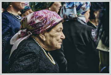 scena rodzajowa - Poziomy, barwny kadr fotograficzny przedstawiający na pierwszym planie postać starszej kobiety w czarnej sukience, białych perłach na szyi oraz wzorzystej chustce w odcieniach fioletu na głowie. Kobieta patrzy przed siebie, na drugim planie widać grupę podobnie ubranych kobiet, których postaci zostały ucięte przez wybrany kadr.
