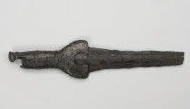 fragment miecza z brązu - Ujęcie z góry; Miecz z brązu zachowany we fragmencie - część głowni oraz rękojeść - o łącznej długości 22,7 cm. Egzemplarz cechuje smukła głownia, na powierzchni której przetrwała miejscami pierwotna lśniąca powłoka barwy ciemnozielonej. Dzięki temu widoczne są po obu stronach ostrza struziny podkreślone delikatnym zdobieniem w postaci drobnych nacięć i łuczków. Podobnym ornamentem, na który składają się pasma linii rytych i niewielkich nacięć, pokryta jest również rękojeść miecza o gładkiej ciemnozielonej powierzchni. Rękojeść jest niekompletna, brakuje jej głowicy – najpewniej tworzyły ją dwie niewielkie woluty, pomiędzy którymi wystawał niewielki pręt.