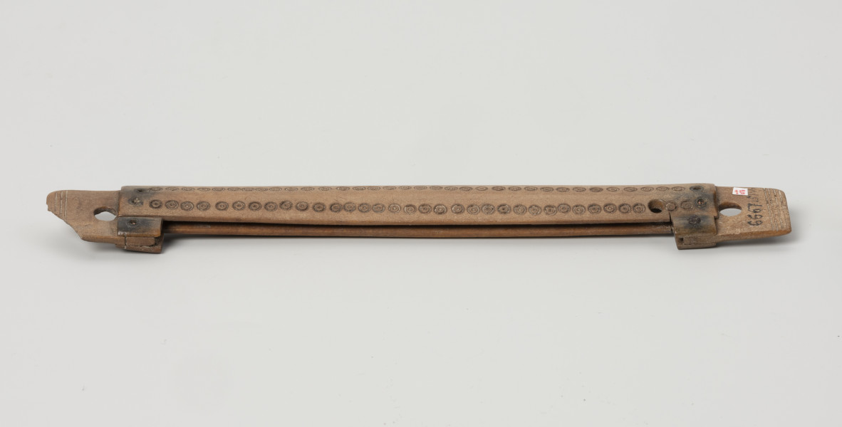 pochewka grzebienia - Ujęcie z tyłu; Obiekt zbudowany został z dwóch okładzin o kształtach wydłużonych prostokątów o prostych bokach, okładziny, ułożone równolegle względem siebie, umocowane są do dwóch bocznych płytek za pomocą nitów wykonanych ze stopu miedzi, po dwa nity na każdą płytkę. Boczne płytki częściowo wchodzą w odstęp pomiędzy okładzinami, wyznaczając w ten sposób szerokość przestrzeni na grzebień, a częściowo wystają poza ich końce. Jedna ich krawędź położona jest w tej samej linii co krawędź okładzin, a druga nieco wystaje. Na tych wystających odcinkach umocowano dodatkowo krótkie fragmenty kolejnych okładzin, zrównanych z jednej strony z okładzinami głównymi, a z drugiej z krawędziami wewnętrznymi płytek. Płytki posiadają koliste otwory do umocowania grzebienia w pochewce, a ich zakończenia zdobione są czterema nacięciami biegnącymi przez całą szerokość. Okładziny główne ozdobiono przy krawędziach dwoma rzędami kolistych oczek z zaznaczonymi środkami. Całość została wypolerowana.