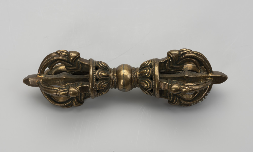 wadżra - ujęcie z przodu; Mały, wykonany z brązu, przypominający berło przedmiot w kształcie kokardy lub ósemki. W jego centrum znajduje się kula - uchwyt berła. Po jej obu stronach znajdują się wyrastające w przeciwnych kierunkach stożkowe zgrubienia stylizowane na kwiaty lotosu z ośmioma płatkami. Od każdego z nich odchodzą cztery, łukowate ramiona zbiegające się wokół centralnej szpicy przebiegającej przez całą oś obiektu. Ramiona bogato zdobione w formie ozdoby przypominającej podwinięty liść przedstawiającej wystylizowaną głową potwora zwanego makarą.