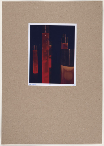 fotografia - Barwna odbitka fotograficzna przedstawiająca pięć wysokich, cylindrycznych kształtów ustawionych w nieregularnym skupisku. Są to walce zakończone połyskliwymi, ciemnymi nasadkami, z przeźroczystego materiału o częściowo transparentnym wypełnieniu w nasyconych barwach (czerwono-pomarańczowych). W środku widoczne są pęcherze powietrza.  Elementy kontrastują z bardzo ciemnym tłem. Odbitka fotograficzna została wykonana z wąską, białą ramką.