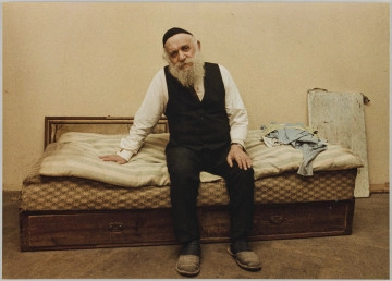 fotografia - Zdjęcie przedstawia starszego mężczyznę z siwą brodą siedzącego na łóżku w mieszkaniu. Ma na sobie czarne spodnie, białą koszulę, czarną kamizelkę i czarną jarmułkę. Jedną rękę opiera na pościeli, drugą na kolanie. Obok niego, na pościeli leżą części odzieży. Jego głowa jest lekko przekrzywiona do boku. Patrzy w obiektyw.