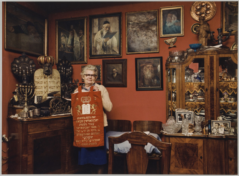 fotografia - Zdjęcie przedstawia starszą kobietę trzymającą w rękach sukienkę na Torę. Kobieta stoi w pomieszczeniu wypełnionym judaikami. Ściany są ozdobione obrazami przedstawiającymi Żydów w różnych sytuacjach. Pólki kredensu (po prawej stronie kadru) oraz blat komody (po lewej stronie kadru) wypełnione są min. świecznikami chanukowymi, kubkami pesachowymi, talerzami sederowymi, a także różnego rodzaju figurkami, naczyniami ze szkła oraz fotografiami. Kobieta stoi przy komodzie. Ma na sobie granatowa spódnicę, kolorowa bluzkę z kołnierzykiem, beżowy sweter oraz okulary. Jej włosy są siwe i zaczesane do tyłu. Sukienka, którą trzyma przed sobą jest w kolorze czerwonym. Ozdobiona złotymi napisami w języku hebrajskim oraz przedstawieniem dwóch lwów trzymających tablice z dziesięciorgiem przykazań w koronie (na górze; nad napisami).