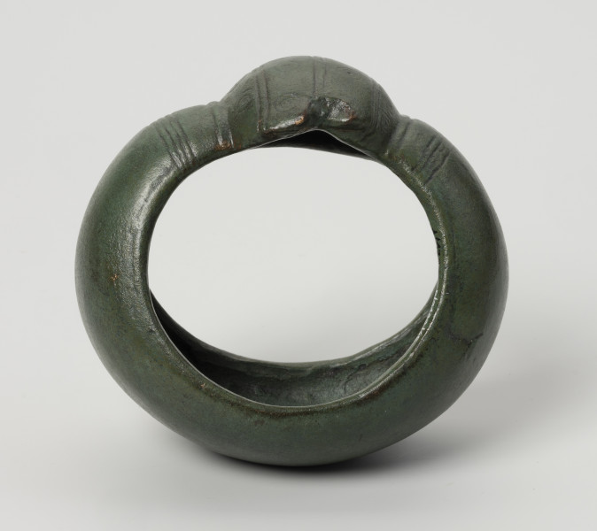 kopia bransolety nerkowatej z Kolkowa - Ujęcie z dołu; Bransoleta o szerokiej obręczy (5,9 cm), silnie wypukła. Jej brzegi są mocno zagięte do środka, co daje C-kształtny przekrój poprzeczny. Oba końce obręczy łączy duże zgrubienie, tzw. nerka. Końce i zgrubienie nerkowate zostały ozdobione pasmami zwielokrotnionych linii (3 pasma przypadają na zgrubienie, a 4 pasma na końce obręczy), które biegną poprzecznie w stosunku do obręczy. Pomiędzy pasmami na zgrubieniu nerkowatym znajduje się tzw. ornament oczkowy (współśrodkowe podwójne okręgi z centralnie usytuowaną kropką-wgłębieniem). Bransoleta ma zielony kolor. Brzeg nerki ma w jednym miejscu zamarkowane uszkodzenie, wzorowane na oryginale bransolety.