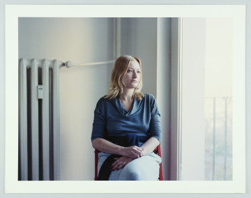 portret fotograficzny - Fotografia barwna skadrowana poziomo przedstawiająca portret kobiety ujętej w 3/4 postaci. Portretowana o jasnych włosach sięgających ramion, ubrana jest w ciemnoniebieską bluzkę o długich rękawach zsuniętych pod łokcie i jasnoniebieskie spodnie. Kobieta siedzi na czerwonym krześle (widać jedynie zarys oparcia) we wnętrzu na tle narożnika białej ściany i dużego okna, trzyma ręce skrzyżowane na kolanach, patrzy w bok. Na ścianie, po lewej stronie postaci jest widoczny duży kaloryfer żeberkowy.  Za oknem widać niewyraźne zarysy krajobrazu.