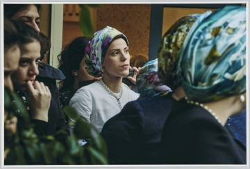 scena rodzajowa - Poziomy, barwny kadr fotograficzny przedstawiający w centralnej części postać kobiety (widocznej po popiersia) w białej sukience, białych perłach na szyi i jasnej, kwiecistej chustce na głowie. Kobieta patrzy w dal, lewą ręką trzyma przy uchu prosty telefon komórkowy. Kobieta stoi w pomieszczeniu, w grupie innych kobiet i dziewcząt.