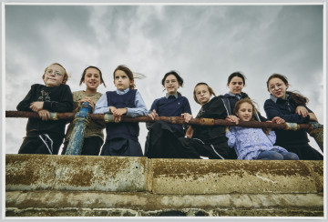 scena rodzajowa - Poziomy, barwny kadr fotograficzny przedstawiający zbiorowy portret dzieci – 5 dziewczynek i 3 chłopców. Chłopcy mają charakterystyczne fryzury chasydzkie (krótkie włosy oraz długie, zakręcone pasma nad uszami), 4 dziewczynki są ubrane w podobne stroje – niebieskie bluzki z kołnierzykami, granatowe spódnice i granatowe swetry (mundurki szkolne), jedna dziewczynka ma niebieską sukienkę w białe drobne kwiatki i szare rajstopy. Dzieci opierają się o zardzewiałą barierkę przymocowaną do betonu, na którym też są ślady rdzy. Fotografia została wykonana od dołu.