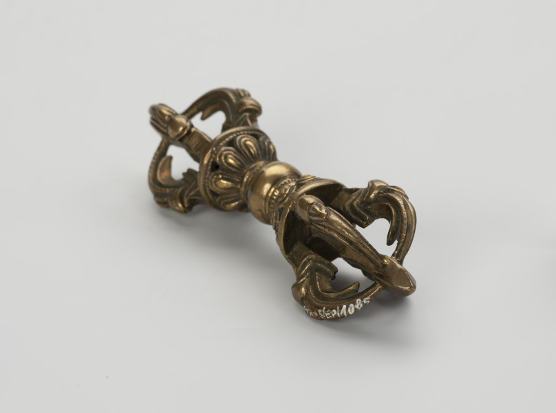 wadżra - ujęcie przodu ze skosu z prawej; Mały, wykonany z brązu, przypominający berło przedmiot w kształcie kokardy lub ósemki. W jego centrum znajduje się kula - uchwyt berła. Po jej obu stronach znajdują się wyrastające w przeciwnych kierunkach stożkowe zgrubienia stylizowane na kwiaty lotosu z ośmioma płatkami. Od każdego z nich odchodzą cztery, łukowate ramiona zbiegające się wokół centralnej szpicy przebiegającej przez całą oś obiektu. Ramiona bogato zdobione w formie ozdoby przypominającej podwinięty liść przedstawiającej wystylizowaną głową potwora zwanego makarą.