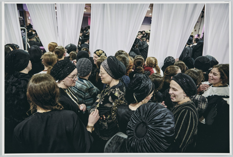 scena rodzajowa - Na pierwszym planie poziomego, barwnego kadru fotograficznego widać dużą grupę, stojących, rozmawiających ze sobą kobiet. Cześć z nich patrzy za białe zasłony, przedzielające pomieszczenie, w którym są na dwie części. Kobiety są ubrane w czarne, stroje, częściowo z elementami dekoracyjnymi lub wzorami w innych kolorach. Niektóre z nich na głowach noszą czepki. Za zasłonami, które wiszą od sufitu w kilku częściach, między którymi są prześwity, widać w bardzo ograniczonym stopniu dalszą część pomieszczenia wypełnioną tłumem mężczyzn w czarnych strojach, kapeluszach lub okrągłych nakryciach głowy (jarmułkach).