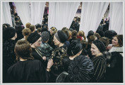 scena rodzajowa - Na pierwszym planie poziomego, barwnego kadru fotograficznego widać dużą grupę, stojących, rozmawiających ze sobą kobiet. Cześć z nich patrzy za białe zasłony, przedzielające pomieszczenie, w którym są na dwie części. Kobiety są ubrane w czarne, stroje, częściowo z elementami dekoracyjnymi lub wzorami w innych kolorach. Niektóre z nich na głowach noszą czepki. Za zasłonami, które wiszą od sufitu w kilku częściach, między którymi są prześwity, widać w bardzo ograniczonym stopniu dalszą część pomieszczenia wypełnioną tłumem mężczyzn w czarnych strojach, kapeluszach lub okrągłych nakryciach głowy (jarmułkach).