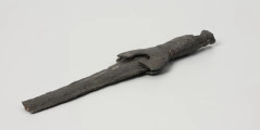 fragment miecza z brązu - Ujęcie z przodu; Miecz z brązu zachowany we fragmencie - część głowni oraz rękojeść - o łącznej długości 22,7 cm. Egzemplarz cechuje smukła głownia, na powierzchni której przetrwała miejscami pierwotna lśniąca powłoka barwy ciemnozielonej. Dzięki temu widoczne są po obu stronach ostrza struziny podkreślone delikatnym zdobieniem w postaci drobnych nacięć i łuczków. Podobnym ornamentem, na który składają się pasma linii rytych i niewielkich nacięć, pokryta jest również rękojeść miecza o gładkiej ciemnozielonej powierzchni. Rękojeść jest niekompletna, brakuje jej głowicy – najpewniej tworzyły ją dwie niewielkie woluty, pomiędzy którymi wystawał niewielki pręt.