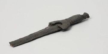 fragment miecza z brązu - Ujęcie z przodu; Miecz z brązu zachowany we fragmencie - część głowni oraz rękojeść - o łącznej długości 22,7 cm. Egzemplarz cechuje smukła głownia, na powierzchni której przetrwała miejscami pierwotna lśniąca powłoka barwy ciemnozielonej. Dzięki temu widoczne są po obu stronach ostrza struziny podkreślone delikatnym zdobieniem w postaci drobnych nacięć i łuczków. Podobnym ornamentem, na który składają się pasma linii rytych i niewielkich nacięć, pokryta jest również rękojeść miecza o gładkiej ciemnozielonej powierzchni. Rękojeść jest niekompletna, brakuje jej głowicy – najpewniej tworzyły ją dwie niewielkie woluty, pomiędzy którymi wystawał niewielki pręt.