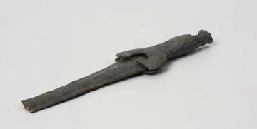 fragment miecza z brązu - Ujęcie z przodu; Miecz z brązu zachowany we fragmencie - część głowni oraz rękojeść - o łącznej długości 22,7 cm. Egzemplarz cechuje smukła głownia, na powierzchni której przetrwała miejscami pierwotna lśniąca powłoka barwy ciemnozielonej. Dzięki temu widoczne są po obu stronach ostrza struziny podkreślone delikatnym zdobieniem w postaci drobnych nacięć i łuczków. Podobnym ornamentem, na który składają się pasma linii rytych i niewielkich nacięć, pokryta jest również rękojeść miecza o gładkiej ciemnozielonej powierzchni. Rękojeść jest niekompletna, brakuje jej głowicy – najpewniej tworzyły ją dwie niewielkie woluty, pomiędzy którymi wystawał niewielki pręt.