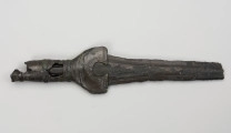 fragment miecza z brązu - Ujęcie z dołu; Miecz z brązu zachowany we fragmencie - część głowni oraz rękojeść - o łącznej długości 22,7 cm. Egzemplarz cechuje smukła głownia, na powierzchni której przetrwała miejscami pierwotna lśniąca powłoka barwy ciemnozielonej. Dzięki temu widoczne są po obu stronach ostrza struziny podkreślone delikatnym zdobieniem w postaci drobnych nacięć i łuczków. Podobnym ornamentem, na który składają się pasma linii rytych i niewielkich nacięć, pokryta jest również rękojeść miecza o gładkiej ciemnozielonej powierzchni. Rękojeść jest niekompletna, brakuje jej głowicy – najpewniej tworzyły ją dwie niewielkie woluty, pomiędzy którymi wystawał niewielki pręt.