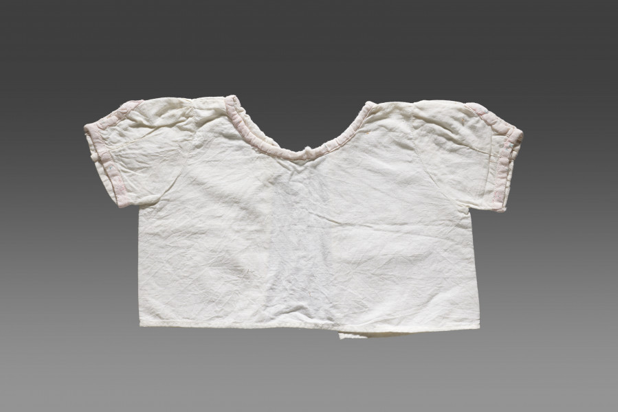 Kaftanik dziecięcy wykonany z cienkiej tkaniny bawełnianej w kolorze białym/écru. Kaftanik o prostym kroju i krótkim rękawku. Rękawy oraz linia dekoltu (brak kołnierzyka) wykończone jasnoróżową tasiemką z widocznym nadrukiem floralnym. Kaftanik wiązany pod szyją na białą tasiemkę. 