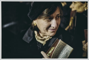 scena rodzajowa - Fotograficzny portret kobiety w średnim wieku z ciemnymi włosami, ubranej w czarny strój, z czarnym nakryciem głowy, z jasną apaszką/kołnierzykiem pod szyją oraz z ozdobnymi złotymi kolczykami. Kobieta ma głowę i wzrok skierowane w prawą stronę (widza). W ręku, na wysokości szyi, trzyma tekst po hebrajsku. Ostrość ustawiona została na twarz portretowanej, tak, że szczegóły stroju oraz tło są mocno niewyraźne – jedynie a prawym górnym rogu widać twarz kobiety.