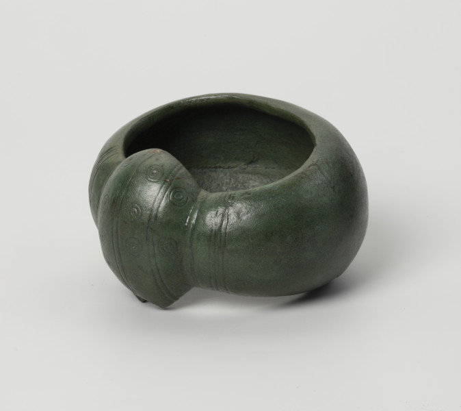 kopia bransolety nerkowatej z Kolkowa - Ujęcie przodu ze skosu z prawej; Bransoleta o szerokiej obręczy (5,9 cm), silnie wypukła. Jej brzegi są mocno zagięte do środka, co daje C-kształtny przekrój poprzeczny. Oba końce obręczy łączy duże zgrubienie, tzw. nerka. Końce i zgrubienie nerkowate zostały ozdobione pasmami zwielokrotnionych linii (3 pasma przypadają na zgrubienie, a 4 pasma na końce obręczy), które biegną poprzecznie w stosunku do obręczy. Pomiędzy pasmami na zgrubieniu nerkowatym znajduje się tzw. ornament oczkowy (współśrodkowe podwójne okręgi z centralnie usytuowaną kropką-wgłębieniem). Bransoleta ma zielony kolor. Brzeg nerki ma w jednym miejscu zamarkowane uszkodzenie, wzorowane na oryginale bransolety.