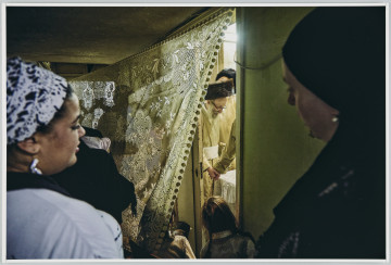 scena rodzajowa - Poziomy, barwny kadr fotograficzny przestawiający na pierwszym, nieostrym planie dwie kobiety, obok postać w czarnej chuście (najpewniej kobieta) trzymająca niemowlę, przed nimi dwie małe dziewczynki zaglądające za zasłonę. Za zasłoną w drugim pomieszczeniu widać fragmentarycznie grupę mężczyzn w odświętnych złotych strojach oraz walcowatych czapkach z futra, stojących wokół stołu. Przez utworzoną szparę widoczne jest, że dwóch z nich trzyma się za ręce.