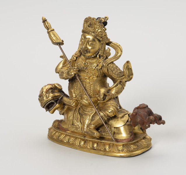 Wajśrawana (Dzambala) - ujęcie przodu ze skosu z prawej; Złota, męska postać siedząca bokiem, z lewą nogą opuszczoną w dół (poza lalitasana), na złotym, ryczącym lwie. W prawej ręce trzyma chorągiew zwycięstwa (osobny, wyciągany element), a w lewej zwierzątko: mangustę  (ichneumon), która wypluwa klejnot z pyszczka. Postać w gniewnej formie, szlachetnie ubrana: nosi diadem, liczne ozdoby z klejnotów, a jej strój przypomina zbroję wysokiej rangi dostojnika/króla. Lwa o bordowych brwiach, sierści na głowie i ogonie przedstawiono w pozycji wpółleżącej - waruje na ugiętych łapach, jest osiodłany, z przodu i z tyłu widoczna uprząż. Obie postacie ustawiono na lotosowej podstawie. W jej centralnym punkcie wydrążono niewielki otwór na szpic sztandaru. Posążek najprawdopodobniej przedstawia bóstwo bogactwa Wajśrawanę wraz z jego atrybutami.