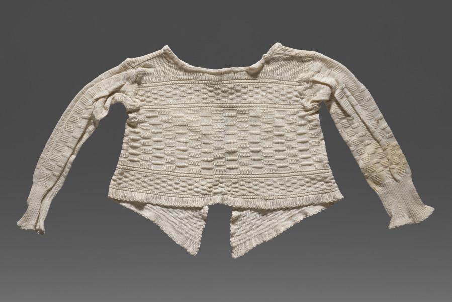 Kaftanik dziecięcy w kolorze écru. Ścieg mieszany, układającymi się w poziome pasy z dominującym motywem szachownicy. W centralnej części wzór jest wyraźnie większy, Wzdół dolnej krawędzi i na wysokości piersi drobniejszy. Poły kaftanika są wydłużone. Kaftanik wiązany pod szyją na taśmę w tym samym kolorze. Rękawy zakończone ściągaczem.