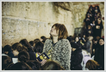 scena rodzajowa - Poziomy, barwny kadr fotograficzny przedstawiający młodą kobietę w stroju europejskim (szarej rozpiętej bluzie z tkaniny przypominającej futerko i bluzce o zwierzęcym wzorze), z rudymi, prostymi włosami do ramion o grzywce opadającej na oczy. Kobieta stoi wyprostowana, z lekko uniesioną ku górze głową – poza wskazuje na głębokie skupienie. Bohaterka fotografii wyróżnia się strojem pozą – jej sylwetka góruje nad dużą grupą kobiet stojących w około tuż przy wysokim murze. Ostrość skierowana jest na kobietę, sylwetki pozostałych oraz otoczenia są rozmyte.