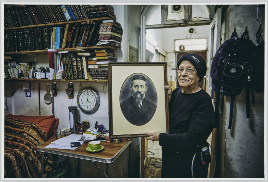 scena rodzajowa - Poziomy, barwny kadr fotograficzny przedstawiający w centralnej części kadru starszą kobietę ubraną w czarny strój i czarne nakrycie głowy. Kobieta stoi przodem do widza i trzyma w geście prezentacji fotograficzny archiwalny portret mężczyzny z brodą w owalnym nakryciu głowy (kipie) oraz marynarce. Fotografia jest oprawiona w passe-partout oraz ramę. Po lewej stronie kadru widoczne jest stojące w narożniku pomieszczenia, zakryte pasiastą narzutą, łóżko, nad nim wiszą 3 rzędy półek wypełnione szczelnie książkami. N pierwszej półce od dołu stoją także buteleczki oraz kubek. Pod półkami wiszą różne drobne przedmioty m.in. koraliki, zegarek. Obok lóżka stoi składany, turystyczny stolik, na nim leżą kartki, książki, stoją naczynia oraz lampka. Nad stolikiem wisi duży, okrągły zegar. Po prawej stronie kadru jest biała ściana, na niej rząd 3 pojedynczych wieszaków, na których wiszą czarne plecami. Za plecami kobiety, w głąb kadru widoczne są otwarte dwuskrzydłowe, przeszklone drzwi, za nimi dalsze pomieszczenie.