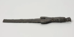 fragment miecza z brązu - Ujęcie z boku; Miecz z brązu zachowany we fragmencie - część głowni oraz rękojeść - o łącznej długości 22,7 cm. Egzemplarz cechuje smukła głownia, na powierzchni której przetrwała miejscami pierwotna lśniąca powłoka barwy ciemnozielonej. Dzięki temu widoczne są po obu stronach ostrza struziny podkreślone delikatnym zdobieniem w postaci drobnych nacięć i łuczków. Podobnym ornamentem, na który składają się pasma linii rytych i niewielkich nacięć, pokryta jest również rękojeść miecza o gładkiej ciemnozielonej powierzchni. Rękojeść jest niekompletna, brakuje jej głowicy – najpewniej tworzyły ją dwie niewielkie woluty, pomiędzy którymi wystawał niewielki pręt.