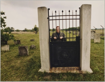 fotografia - Zdjęcie przedstawia mężczyznę stojącego za zamkniętą bramą. Brama składa się z gładkiej metalowej płyty sięgającej połowy wysokości, pionowych prętów o ozdobnych zwieńczeniach (od połowy wysokości do góry) oraz prętów poziomych (dwóch na górze i jednego na dole). Mężczyzna opiera się rękami o jeden z poziomych prętów bramy (umieszczony najbliżej płyty). Patrzy przed siebie. Ma na sobie ciemną kurtkę oraz jarmułkę. Brama jest umocowana pomiędzy dwoma betonowymi słupami. Brakuje jednak reszty ogrodzenia. Za bramą widoczna łąka oraz wyrastające z ziemi pojedyncze macewy i drzewa. 