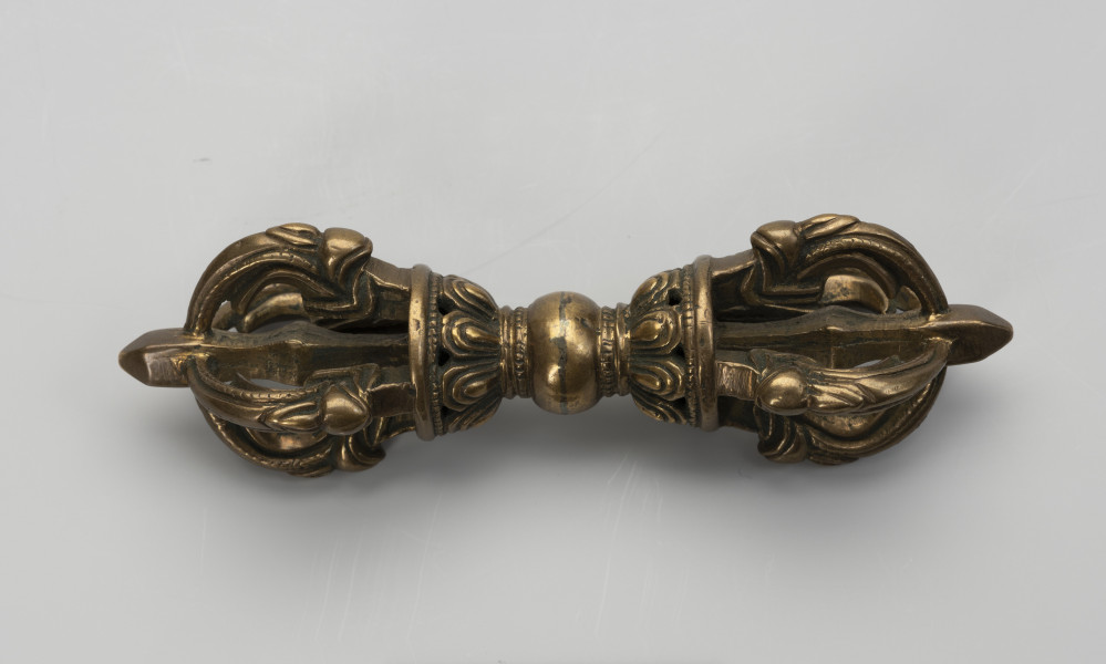 wadżra - ujęcie z przodu; Mały, wykonany z brązu, przypominający berło przedmiot w kształcie kokardy lub ósemki. W jego centrum znajduje się kula - uchwyt berła. Po jej obu stronach znajdują się wyrastające w przeciwnych kierunkach stożkowe zgrubienia stylizowane na kwiaty lotosu z ośmioma płatkami. Od każdego z nich odchodzą cztery, łukowate ramiona zbiegające się wokół centralnej szpicy przebiegającej przez całą oś obiektu. Ramiona bogato zdobione w formie ozdoby przypominającej podwinięty liść przedstawiającej wystylizowaną głową potwora zwanego makarą.