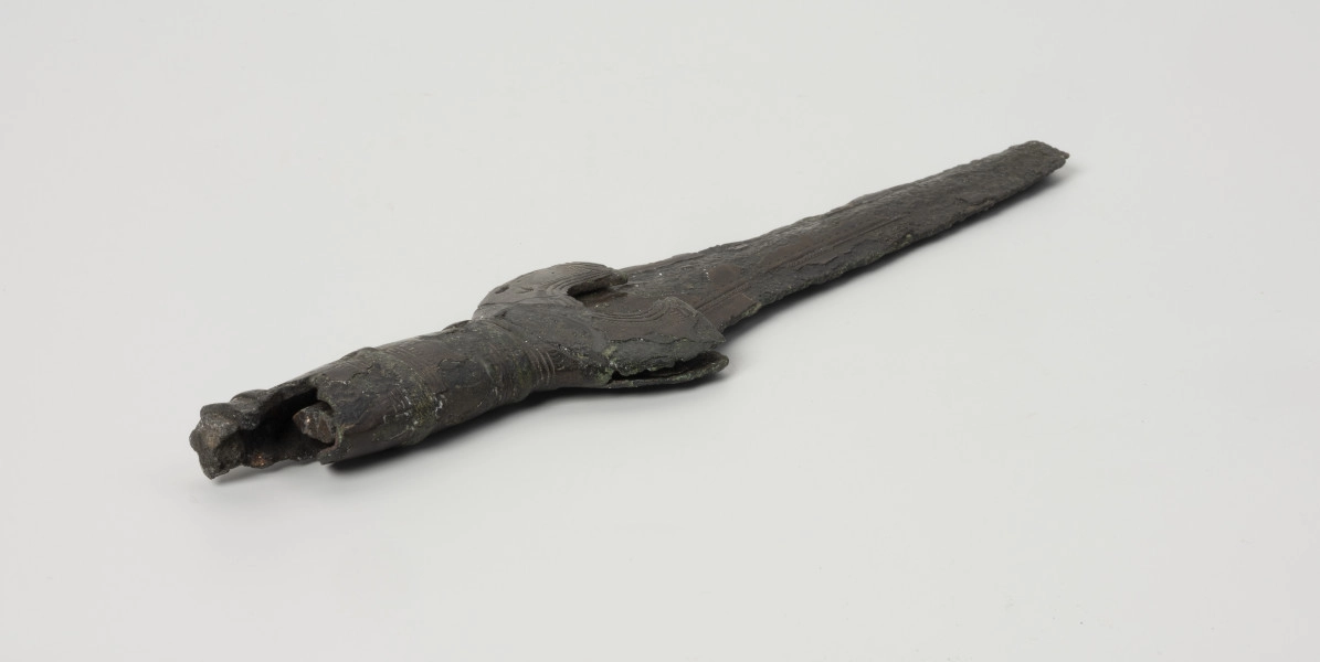 fragment miecza z brązu - Ujęcie z tyłu; Miecz z brązu zachowany we fragmencie - część głowni oraz rękojeść - o łącznej długości 22,7 cm. Egzemplarz cechuje smukła głownia, na powierzchni której przetrwała miejscami pierwotna lśniąca powłoka barwy ciemnozielonej. Dzięki temu widoczne są po obu stronach ostrza struziny podkreślone delikatnym zdobieniem w postaci drobnych nacięć i łuczków. Podobnym ornamentem, na który składają się pasma linii rytych i niewielkich nacięć, pokryta jest również rękojeść miecza o gładkiej ciemnozielonej powierzchni. Rękojeść jest niekompletna, brakuje jej głowicy – najpewniej tworzyły ją dwie niewielkie woluty, pomiędzy którymi wystawał niewielki pręt.