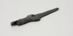 fragment miecza z brązu - Ujęcie z tyłu; Miecz z brązu zachowany we fragmencie - część głowni oraz rękojeść - o łącznej długości 22,7 cm. Egzemplarz cechuje smukła głownia, na powierzchni której przetrwała miejscami pierwotna lśniąca powłoka barwy ciemnozielonej. Dzięki temu widoczne są po obu stronach ostrza struziny podkreślone delikatnym zdobieniem w postaci drobnych nacięć i łuczków. Podobnym ornamentem, na który składają się pasma linii rytych i niewielkich nacięć, pokryta jest również rękojeść miecza o gładkiej ciemnozielonej powierzchni. Rękojeść jest niekompletna, brakuje jej głowicy – najpewniej tworzyły ją dwie niewielkie woluty, pomiędzy którymi wystawał niewielki pręt.