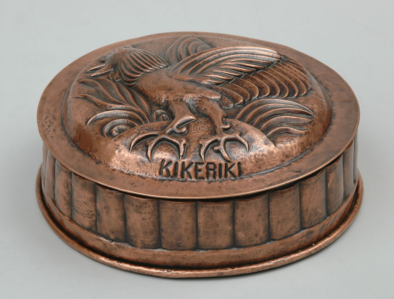 ujęcie 3/4. Naczynie w kształcie walca. Wierzch zdobiony półplastyczną dekoracją i napisem KIKERIKI.