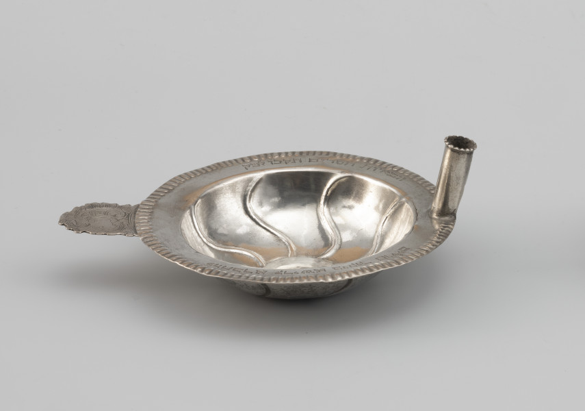 miseczka na datki Chewry Kadiszy - Ujęcie z przodu; Okrągła, głęboka miseczka z lekko wypukłym dnem i płaskim kołnierzem. Kołnierz obwiedziony repusowanym ornamentem sztabkowym. Po lewej stronie miseczki do kołnierza przylutowany poziomo płaski uchwyt o nieregularnym wykroju, zdobiony motywem roccaillowym, po przeciwnej stronie okrągła tulejka, służąca zapewne do mocowania świeczki. Na kołnierzu wygrawerowana inskrypcja fundacyjna w języku hebrajskim z datą.