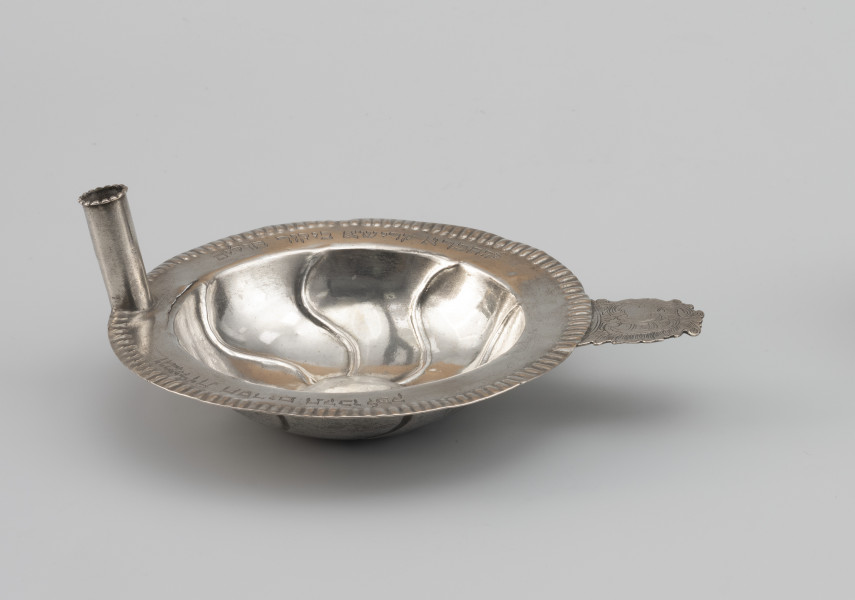 miseczka na datki Chewry Kadiszy - Ujęcie z przodu; Okrągła, głęboka miseczka z lekko wypukłym dnem i płaskim kołnierzem. Kołnierz obwiedziony repusowanym ornamentem sztabkowym. Po prawej stronie miseczki do kołnierza przylutowany poziomo płaski uchwyt o nieregularnym wykroju, zdobiony motywem roccaillowym, po przeciwnej stronie okrągła tulejka, służąca zapewne do mocowania świeczki. Na kołnierzu wygrawerowana inskrypcja fundacyjna w języku hebrajskim z datą.