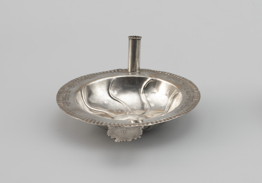 miseczka na datki Chewry Kadiszy - Ujęcie z boku; Okrągła, głęboka miseczka z lekko wypukłym dnem i płaskim kołnierzem. Kołnierz obwiedziony repusowanym ornamentem sztabkowym. Na przodzie miseczki do kołnierza przylutowany poziomo płaski uchwyt o nieregularnym wykroju, zdobiony motywem roccaillowym, po przeciwnej stronie okrągła tulejka, służąca zapewne do mocowania świeczki. Na kołnierzu wygrawerowana inskrypcja fundacyjna w języku hebrajskim z datą.