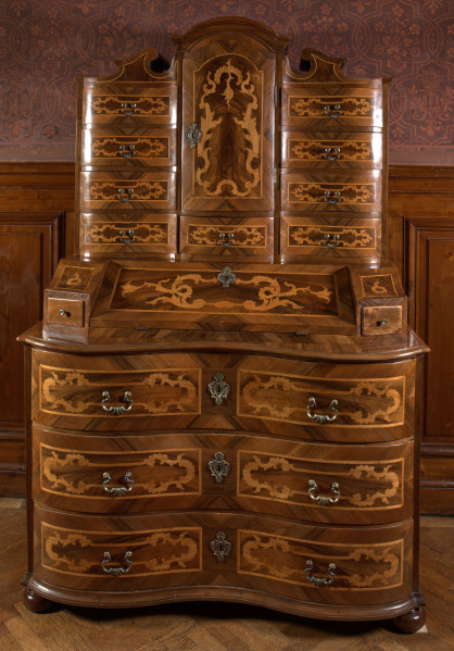 mebel o konstrukcji skrzyniowej, z wybrzuszonym frontem, na czterech kulistych nogach, wykonany z drewna iglastego, odłogowany (okleinowany) orzechem i intarsjowany jesionem (?) w motyw rocaili, część dolną mebla stanowi komoda o trzech szufladach; na niej sekretarzyk z uchylnym blatem-drzwiczkami ustawionym pod kątem i flankowanym dwiema szufladkami, wewnątrz dwa rzędy po cztery mniejsze szufladki; nastawa w formie szafki jednodrzwiowej z flankującymi ją szufladami (po cztery z każdej strony i pod drzwiczkami szafki