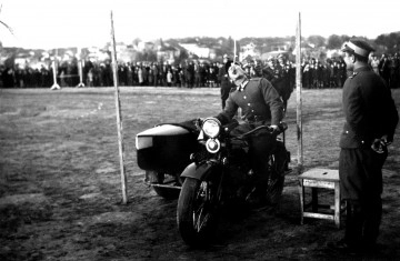 MZŁ-DRM-1901/26 Zdjęcie ukazuje zawody w wyszkoleniu indywidualnym w jeździe na motocyklu. Mężczyzna na motocyklu przejeżdzający pod przeszkodą, z otwartymi ustami próbując chwycić wiszący przedmiot.