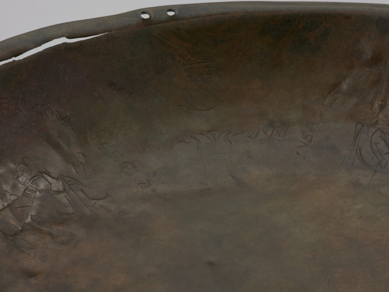 misa z rytem i tekstem - Ujęcie fragmentu obrzeża misay; misa wykonana została ze stopu miedzi, z wąskim, wywiniętym, lekko pogrubionym brzegiem i płaskim dnem. Wewnętrzną powierzchnię ścianki zdobią cztery medaliony ze scenami z życia Heraklesa, otoczone łacińskimi inskrypcjami będącymi cytatami z eklogi przypisywanej Theodolusowi, które zapisano majuskułą romańską. Medaliony przedzielono wizerunkami koronowanych głów męskich.