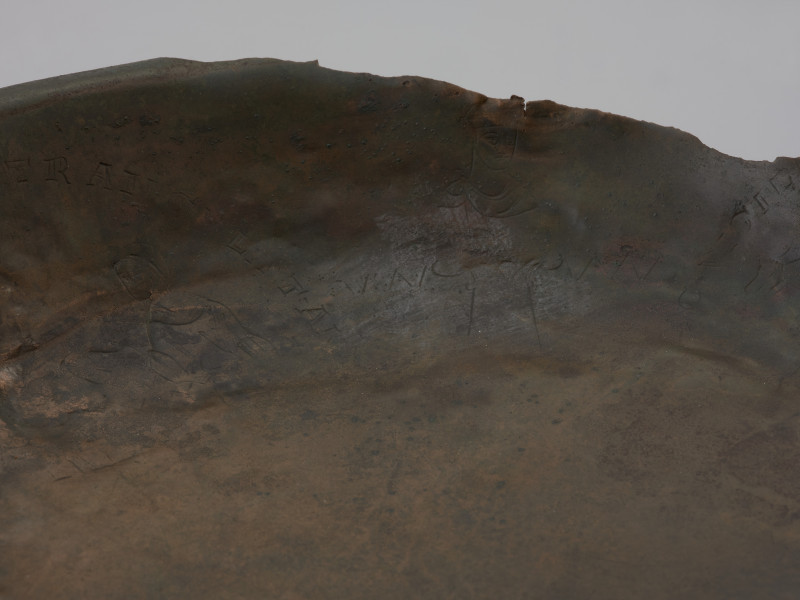 misa z rytem i tekstem - Ujęcie detal; Misa wykonana została ze stopu miedzi, z wąskim, wywiniętym, lekko pogrubionym brzegiem i płaskim dnem. Wewnętrzną powierzchnię ścianki zdobią cztery medaliony ze scenami z życia Heraklesa, otoczone łacińskimi inskrypcjami będącymi cytatami z eklogi przypisywanej Theodolusowi, które zapisano majuskułą romańską. Medaliony przedzielono wizerunkami koronowanych głów męskich.